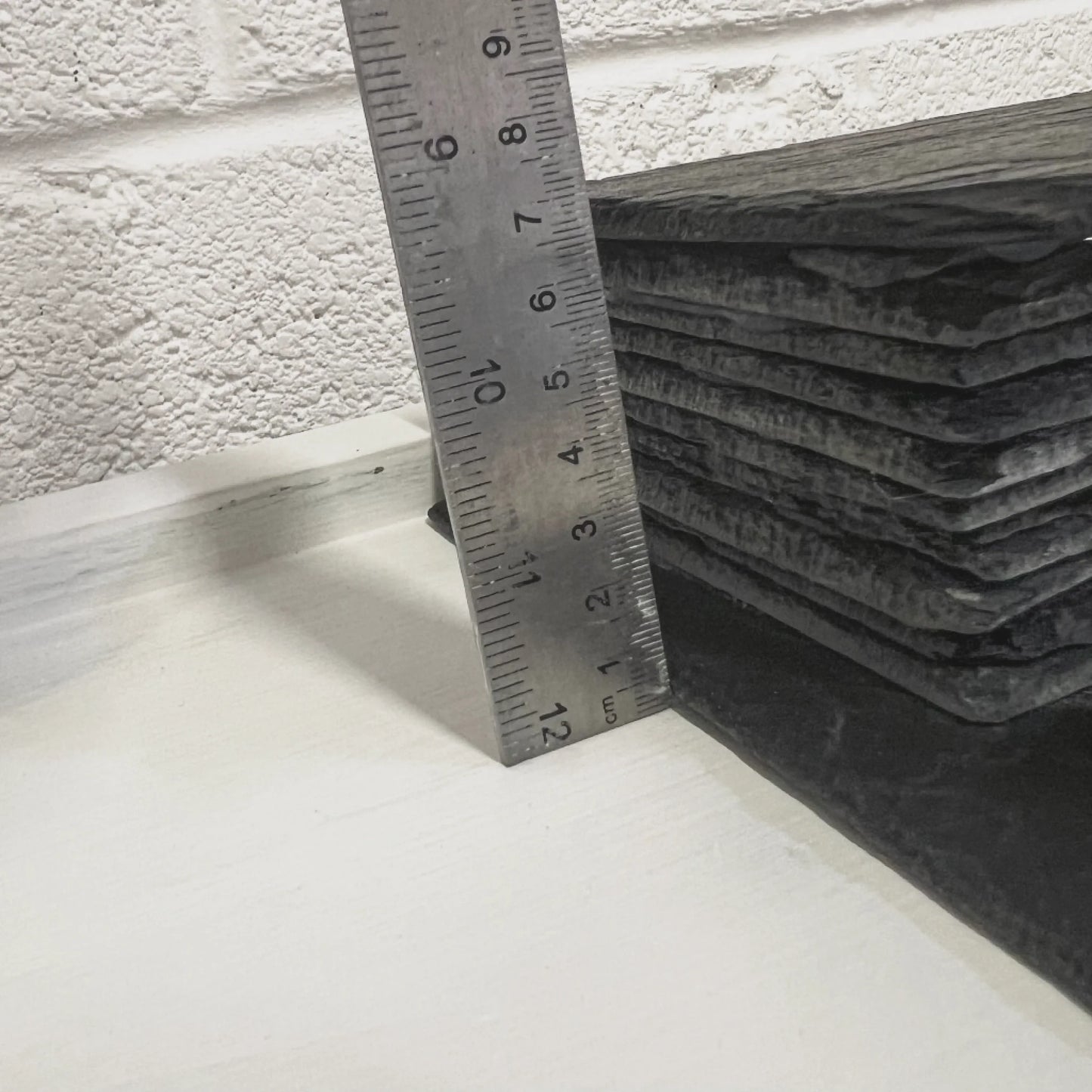 Close-up of handmade black slate reptile hides stacked beside a metal ruler against a white wall.