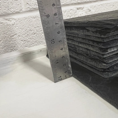 Close-up of handmade black slate reptile hides stacked beside a metal ruler against a white wall.
